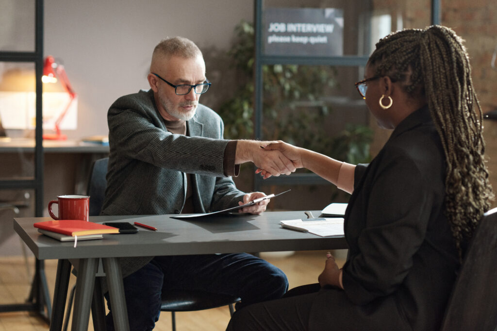 Mature manager shaking hands with young woman while they sitting at table during job interview